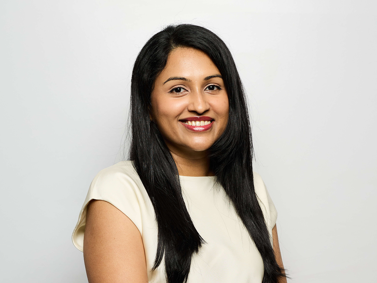 Ms Bharathi Kumaran in a cream blouse smiling with one arm resting on the other against a light grey background